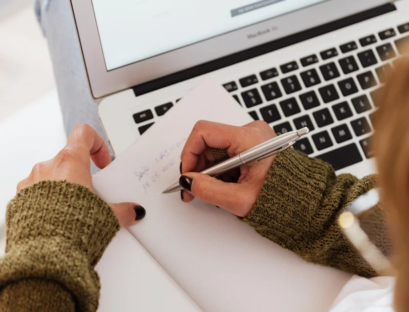 Person writing notes beside laptop.